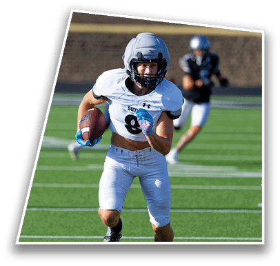 A person wearing a white football uniform and helmet is running with a football while another person in a dark uniform is in the background on a football field. Description generated by AI