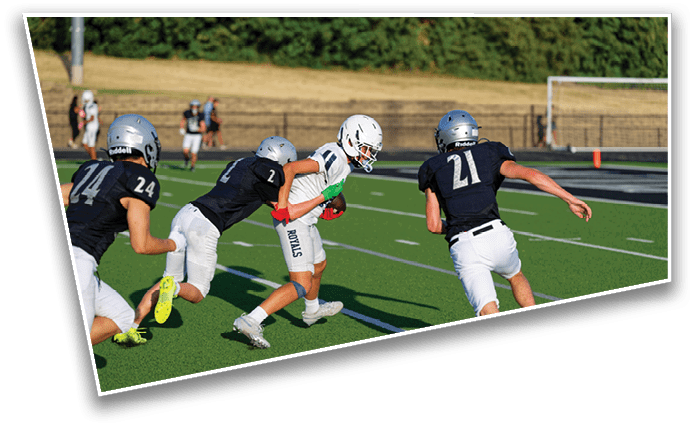People wearing helmets and sports uniforms are playing football on a green field with one person in white running while two others in dark uniforms attempt to tackle. Description generated by AI