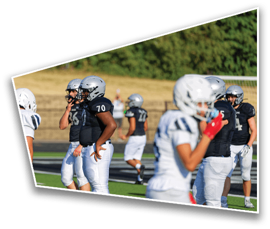 People wearing football helmets and uniforms are standing on a field, with some preparing for play and others walking in the background. Description generated by AI