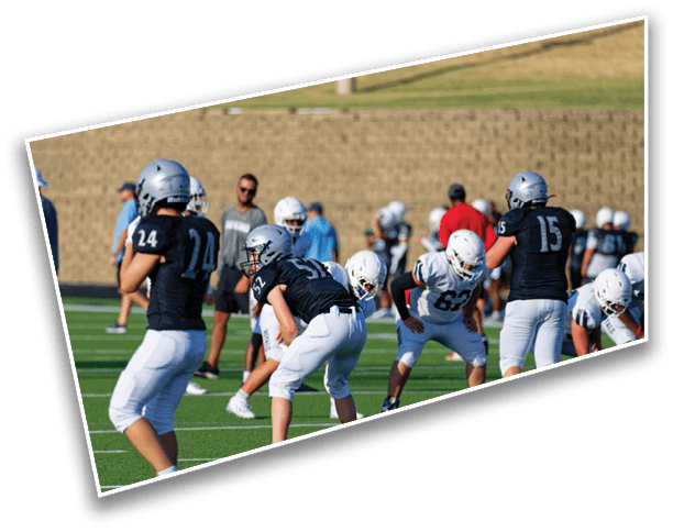 People wearing football helmets and uniforms are positioned on a field preparing for a play during a game or practice. Description generated by AI