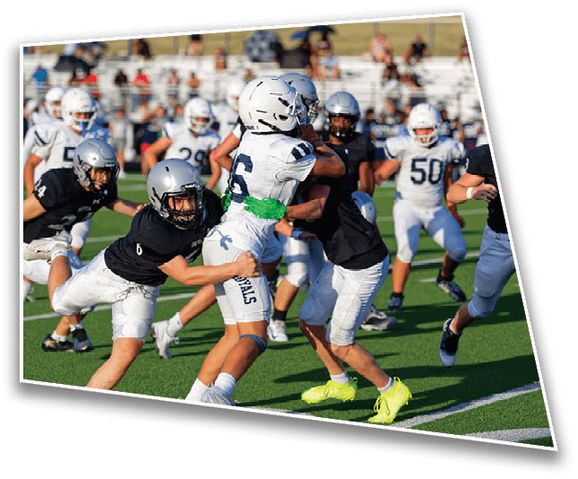 Several people wearing helmets and sports uniforms are actively engaged in a football game on a green field with spectators in the background. Description generated by AI