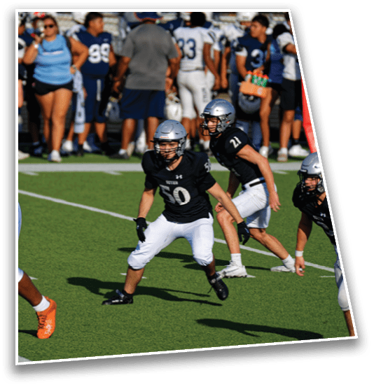 People in football uniforms are actively engaged in a game on a green field with some spectators visible in the background. Description generated by AI