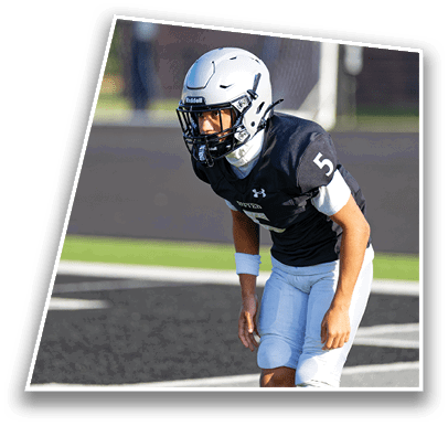 A person wearing a football helmet and uniform is crouched in a ready position on a sports field with a goalpost in the background. Description generated by AI