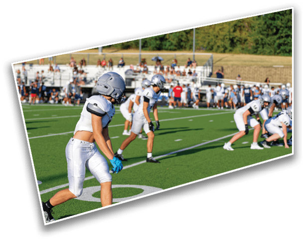 Several people in football uniforms and helmets are positioned on a green field with a crowd seated in bleachers in the background. Description generated by AI