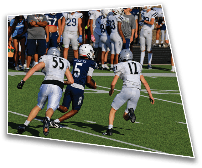 Three football players in action on a green field with one player in a dark uniform running between two players in white uniforms while a group of players and people stand in the background. Description generated by AI
