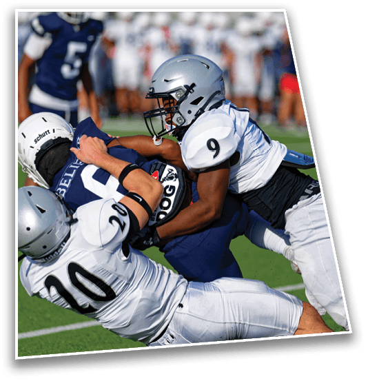 Two people in football uniforms are engaged in a tackle on a green field with blurred players and spectators in the background. Description generated by AI