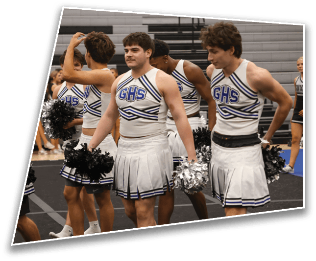 Several people wearing cheerleading uniforms with “GHS" on the front are standing indoors holding pom-poms, with some looking in different directions. Description generated by AI