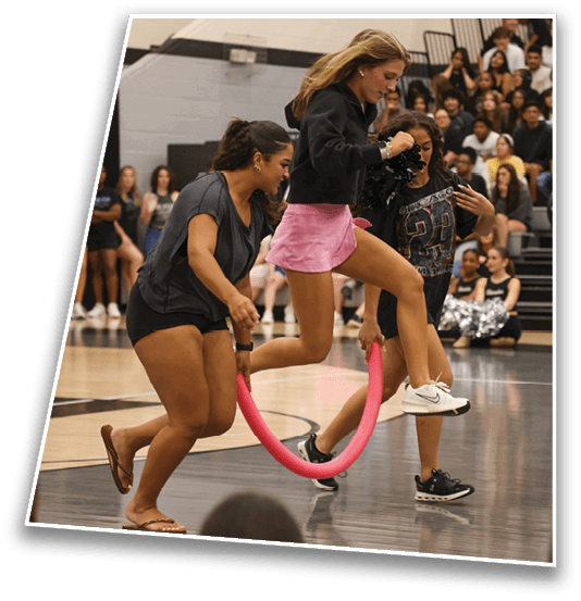 Three people are playing a game indoors where one person is jumping through a pink hoop held by the other two, with an audience seated in the background. Description generated by AI