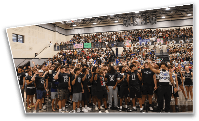 A large group of people wearing matching dark athletic shirts are gathered in a gymnasium with a crowd and band playing in the bleachers behind them. Description generated by AI