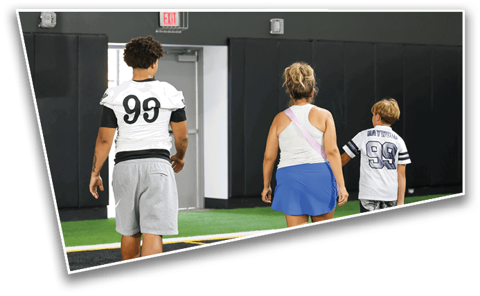 Three people wearing sports jerseys with the number 99 walk away on an indoor turf field toward a door. Description generated by AI