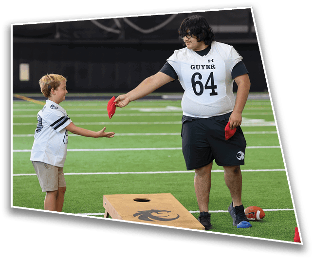 Two people are playing cornhole indoors on a turf field, with one person handing a red bean bag to the other near a wooden cornhole board. Description generated by AI