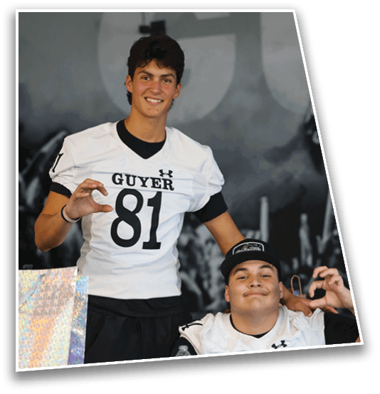 Two people wearing white football jerseys with the word “GUYER" and numbers 81 and 54 pose indoors, with one person standing and the other sitting while holding a water bottle. Description generated by AI