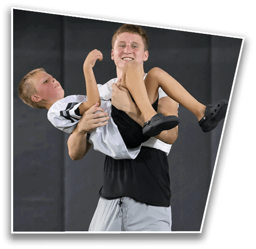 A smiling adult wearing a sleeveless shirt is holding a child dressed in a sports jersey and shorts in their arms against a dark background. Description generated by AI
