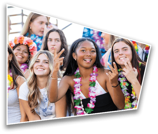 A group of smiling people wearing colorful leis and casual clothing are gathered closely together, with some making peace signs and one person playfully sticking out their tongue. Description generated by AI