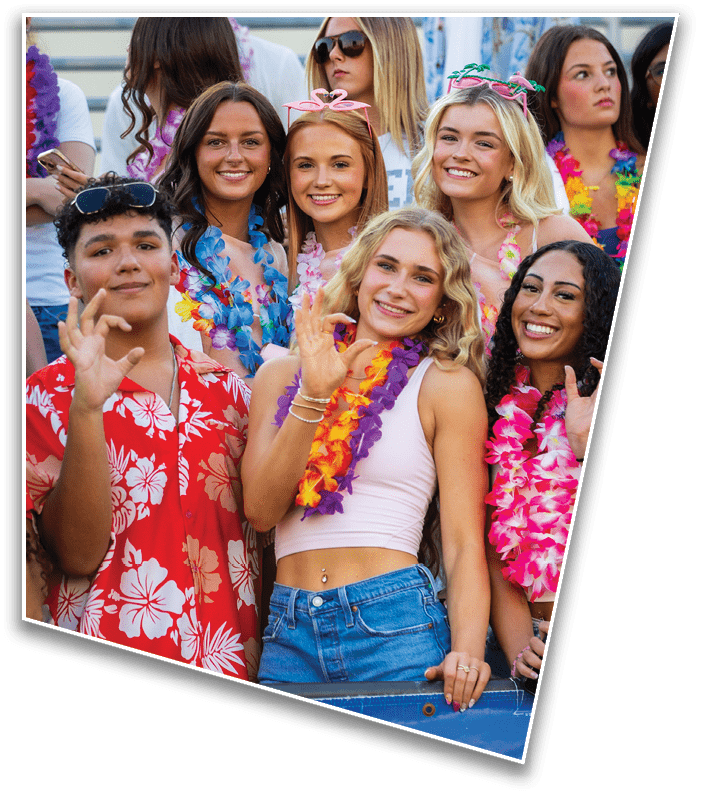 A group of smiling adults wearing colorful leis and festive headbands pose closely together outdoors. Description generated by AI