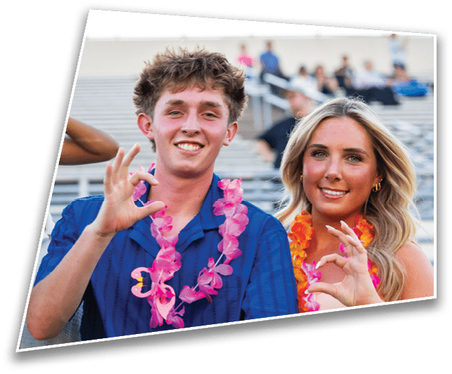 Two adults wearing colorful leis smile and gesture with their hands while sitting in stadium bleachers. Description generated by AI