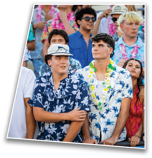 A group of people wearing colorful Hawaiian shirts and leis are standing closely together, appearing to watch an event with focused expressions. Description generated by AI
