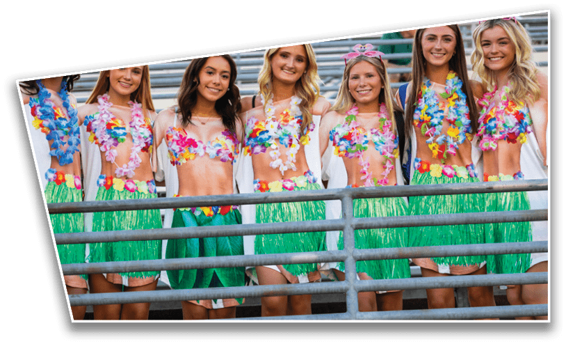 A group of people dressed in colorful Hawaiian-themed outfits with grass skirts and flower leis are standing behind a metal railing, smiling. Description generated by AI