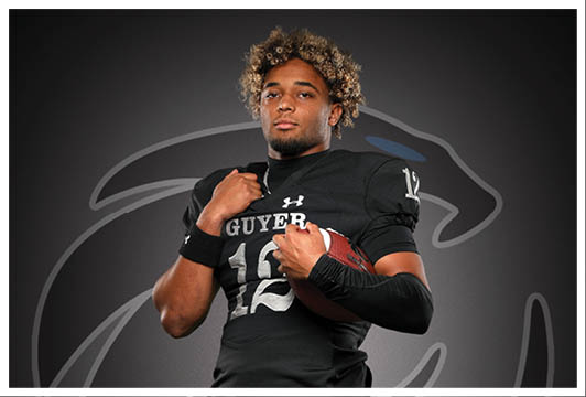 A person with curly hair wearing a black football uniform holding a football against a dark background with a large stylized panther logo. Description generated by AI