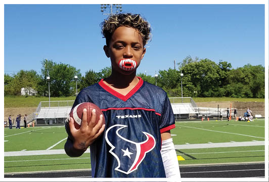 A person with short curly hair wearing a Houston Texans football jersey and a mouthguard is holding a football on a sports field with bleachers and trees in the background. Description generated by AI
