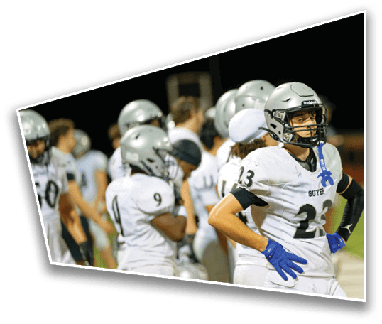 A group of people wearing football helmets and white jerseys are standing together on a field at night, with one person in the foreground wearing number 23 and a blue ribbon on their jersey. Description generated by AI