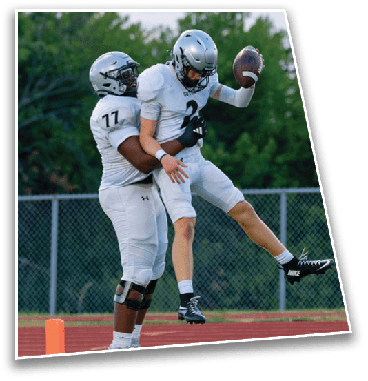 Two people wearing football helmets and white jerseys are engaged in a play on a field with trees and a sunset sky in the background. Description generated by AI