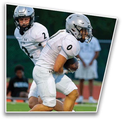Two people in white football uniforms are on a field, with one holding a football and the other positioned behind. Description generated by AI