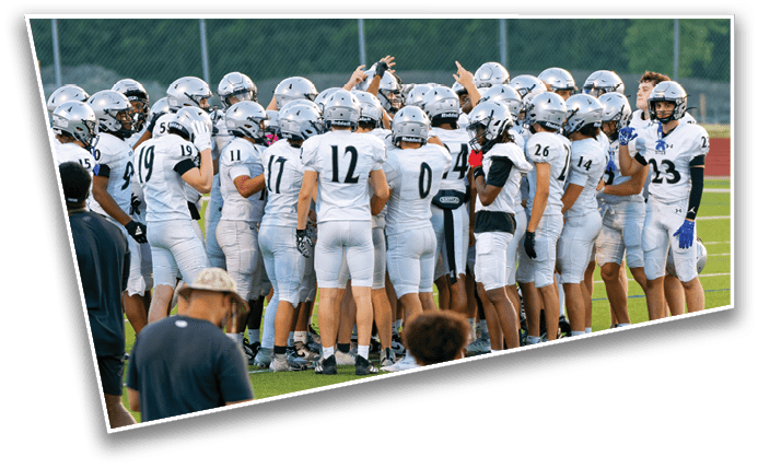 A group of people wearing white football uniforms and helmets are huddled together on a green field with a track and trees in the background. Description generated by AI