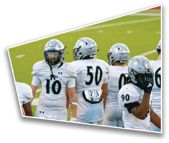 Several people wearing white football uniforms and helmets stand on a green field, with some facing forward and others turned away. Description generated by AI