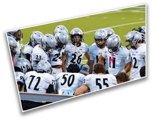 A group of people wearing silver helmets and white sports uniforms are gathered closely together on a green field, appearing to be in a team huddle. Description generated by AI
