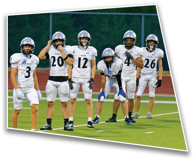 Six people wearing white football uniforms and helmets stand on a green field with a chain-link fence in the background. Description generated by AI
