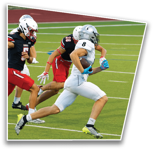 A person in white sports gear is running with a football while being chased by a person in black and red sports gear on a green field. Description generated by AI