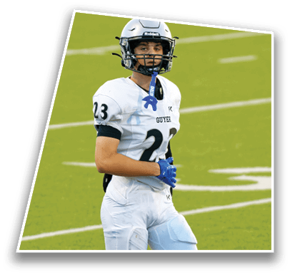 A person wearing a white football jersey with the number 23 and the word Guyer is standing on a field with a helmet and a blue mouthguard hanging from the face mask. Description generated by AI