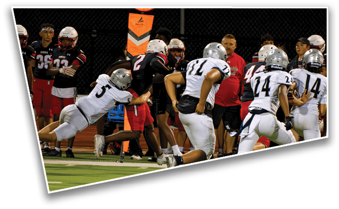 People in football uniforms are actively playing on a field with one player diving to tackle another while others run nearby and coaches or team members stand on the sideline. Description generated by AI
