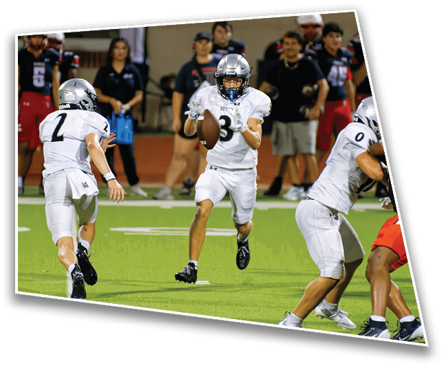 People in football uniforms are actively playing on a field with one person holding a football and others running or preparing to block. Description generated by AI
