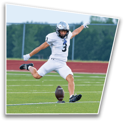 A person in a white football uniform and helmet is kicking a football on a green field with a red track and trees in the background. Description generated by AI