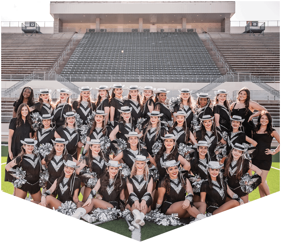A large group of adults dressed in coordinated black and silver dance uniforms with hats and pom-poms pose together on a football field with empty stadium seats in the background. Description generated by AI