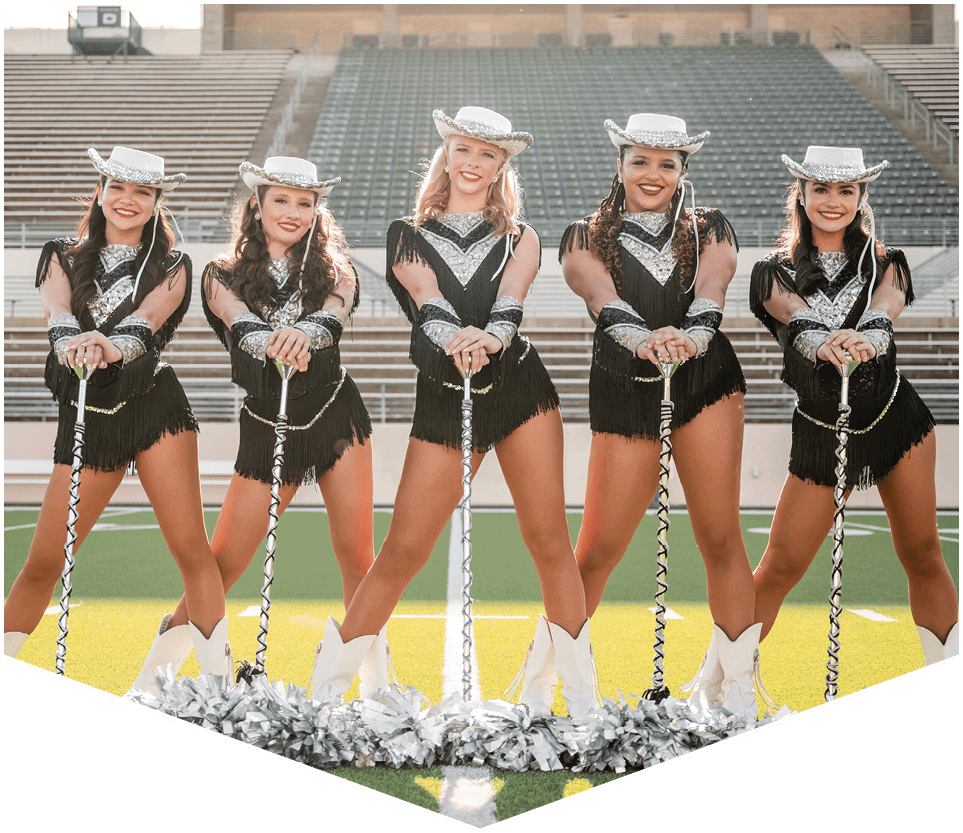 Five adults dressed in coordinated black and silver dance costumes with white cowboy hats and boots stand in a row on a sports field holding batons with silver pom-poms at their feet. Description generated by AI