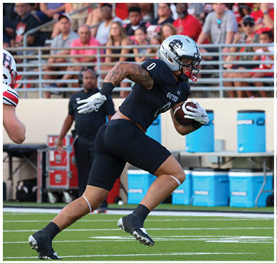 A person wearing a dark sports uniform is running on a football field while holding a football close to their chest. Description generated by AI
