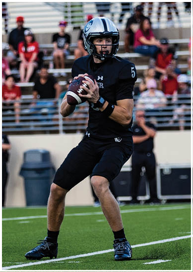 A person wearing a football helmet and black jersey with the number five is holding a football and looking to the side on a field with blurred spectators in the background. Description generated by AI