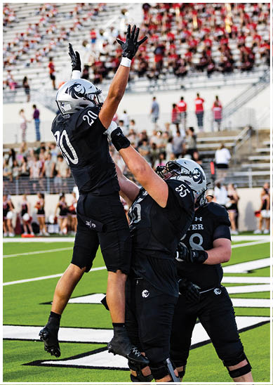 A person wearing a black football uniform with the number 20 and a silver helmet is reaching up with both hands during a game in a stadium with blurred spectators in the background. Description generated by AI