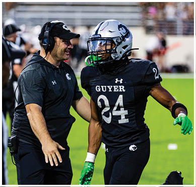 A person wearing a headset and cap is talking to a football player in a helmet and uniform labeled “GUYER" on a sports field. Description generated by AI