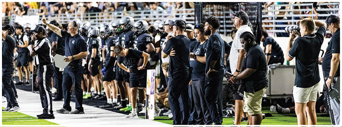A group of people wearing black shirts and helmets stand on a sports field with a large crowd seated in the background. Description generated by AI