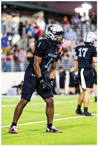 A person wearing a black football uniform and white helmet is on a field with blurred spectators and lights in the background during dusk. Description generated by AI