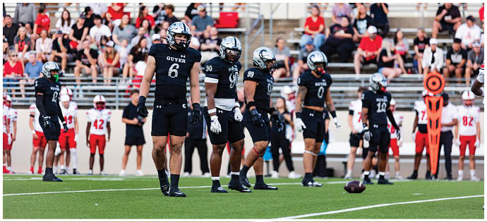 A group of athletes in black football uniforms stand on a field with spectators seated in bleachers behind them. Description generated by AI