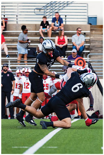 Two football players in black uniforms are tackling a player in a white and red uniform on a green field with spectators in the background. Description generated by AI