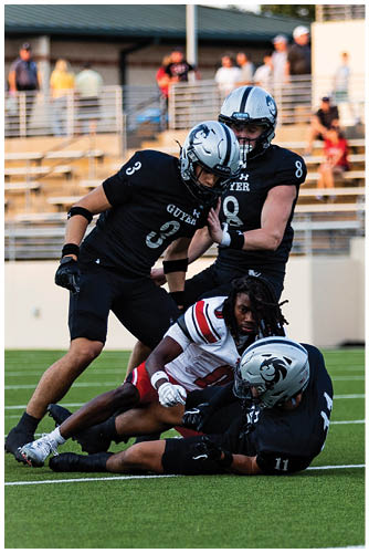 Two people wearing white football helmets with blue stripes are in the foreground with blurred people and a building in the background. Description generated by AI
