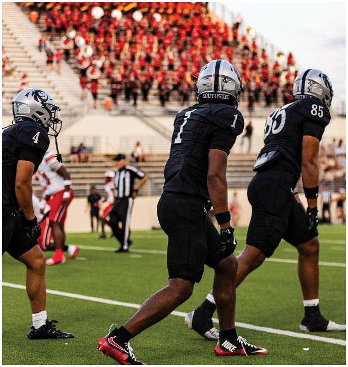 Three people in black football uniforms and helmets are positioned on a field with a crowd and band visible in the background. Description generated by AI
