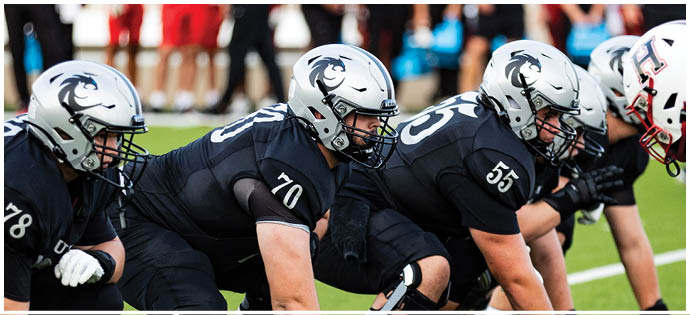 Football players wearing helmets are lined up on the field with coaches and other team members standing in the background. Description generated by AI