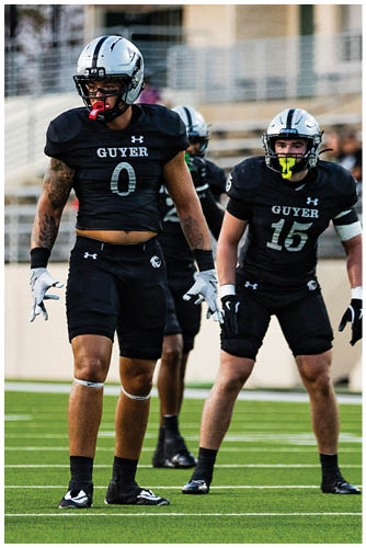 An adult wearing a black football uniform with the number 15 and the word Guyer on the front is on a field with a blurred background of bleachers and another player. Description generated by AI